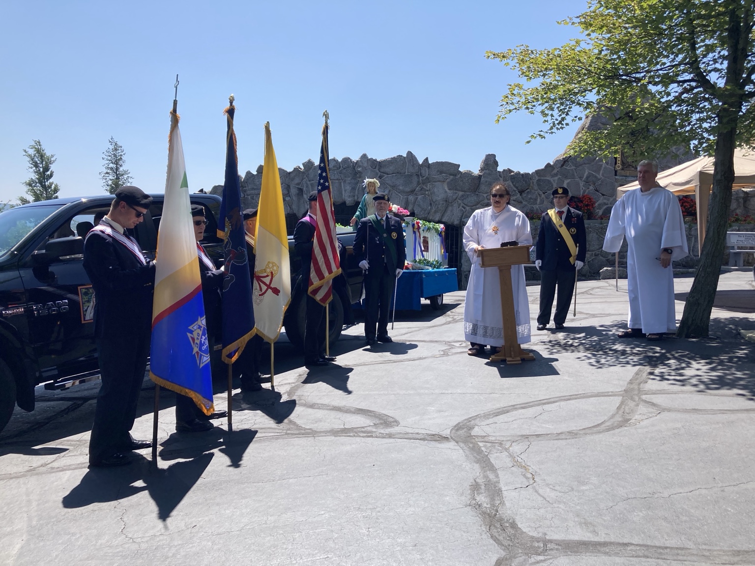 Uniformed flag bearers and clergy beside a flower-decorated statue outdoors.